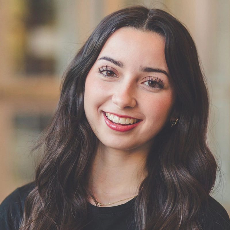 A young woman with long, wavy dark brown hair smiles warmly. She wears a black top, subtle earrings, and a thin gold necklace. The background is softly blurred in neutral tones, keeping the focus on her cheerful expression.
