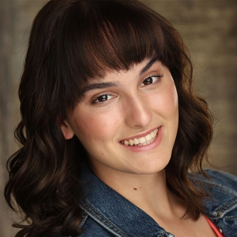 A young woman with wavy dark brown hair and bangs smiles warmly. She wears a denim jacket over a pink top. The background is softly blurred, highlighting her friendly expression and natural makeup.