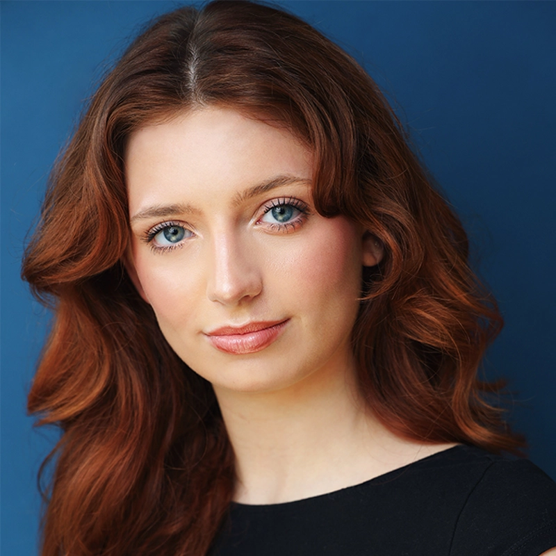 A young woman with wavy auburn hair and blue eyes looks at the camera with a gentle smile. She has fair skin and wears a black top. The background is a solid deep blue, creating a simple, professional portrait style.
