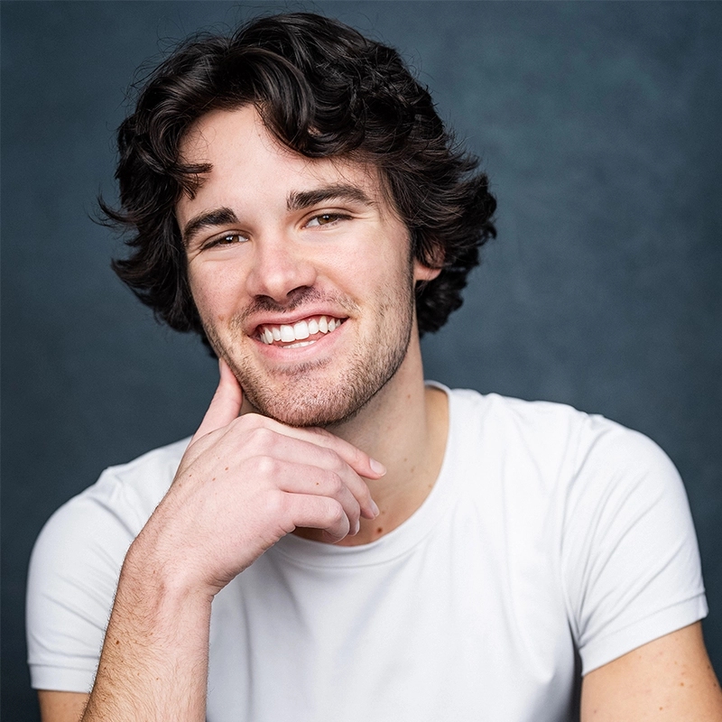A young man with wavy dark hair and light facial hair smiles warmly. He wears a white t-shirt and rests his chin on his hand. The solid, textured dark blue background creates a professional, friendly portrait for Grace Rundberg's profile.