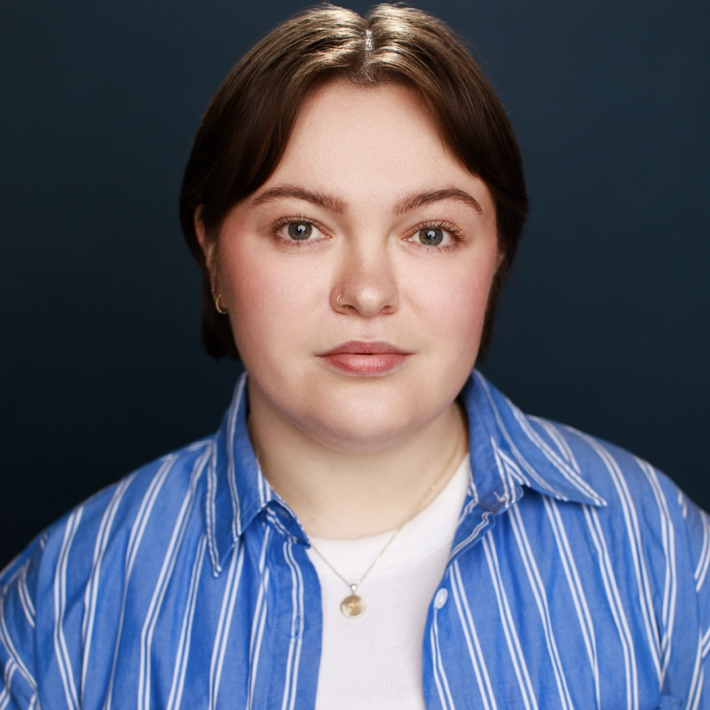 Noelle Johnson, a person with short brown hair and light skin, faces the camera against a dark blue background. They wear a blue-and-white striped shirt over a white top and a gold necklace. Their expression is neutral or slightly smiling.