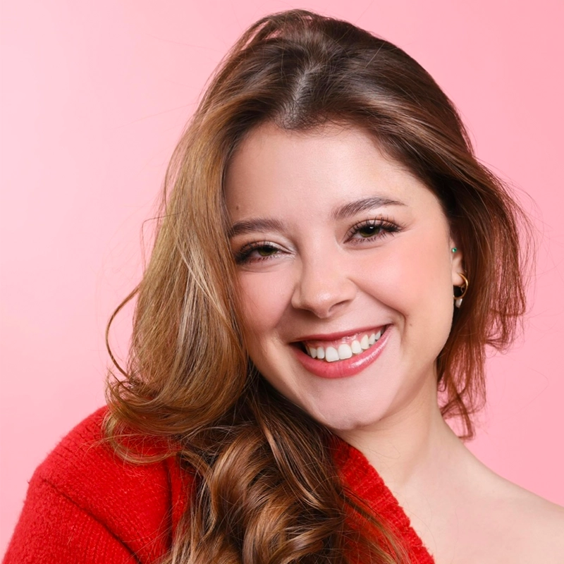 Theresa Landis, a young woman with long, wavy brown hair, smiles brightly at the camera. She wears a red sweater and small earrings, with natural makeup. The solid light pink background creates a cheerful and warm atmosphere.