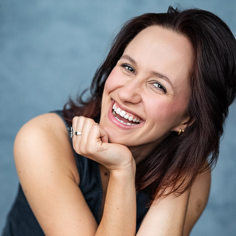 Grace Rundberg, a young woman with shoulder-length brown hair, smiles brightly, showing her teeth. She rests her chin on her hand, wears a sleeveless black top, a silver ring, and small gold hoops. The background is softly blurred blue-gray.