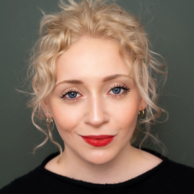 A woman with fair skin, curly blonde hair in a loose updo, and blue eyes is facing forward, smiling softly. She wears bright red lipstick, subtle makeup, gold hoop earrings, and a black top, against a plain, dark gray background.