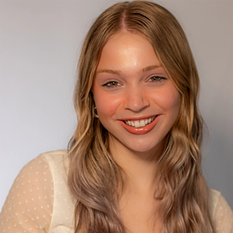 A young woman with long, wavy blonde hair smiles at the camera. Theresa Landis is wearing a sheer, light-colored top with small polka dots. The plain, light gray background and soft lighting highlight her natural features.