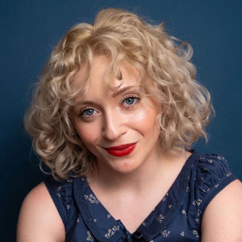 A woman with light skin and short, curly blonde hair smiles softly. She is wearing a navy blue sleeveless top with a subtle pattern and bright red lipstick. The background is a solid dark blue color.