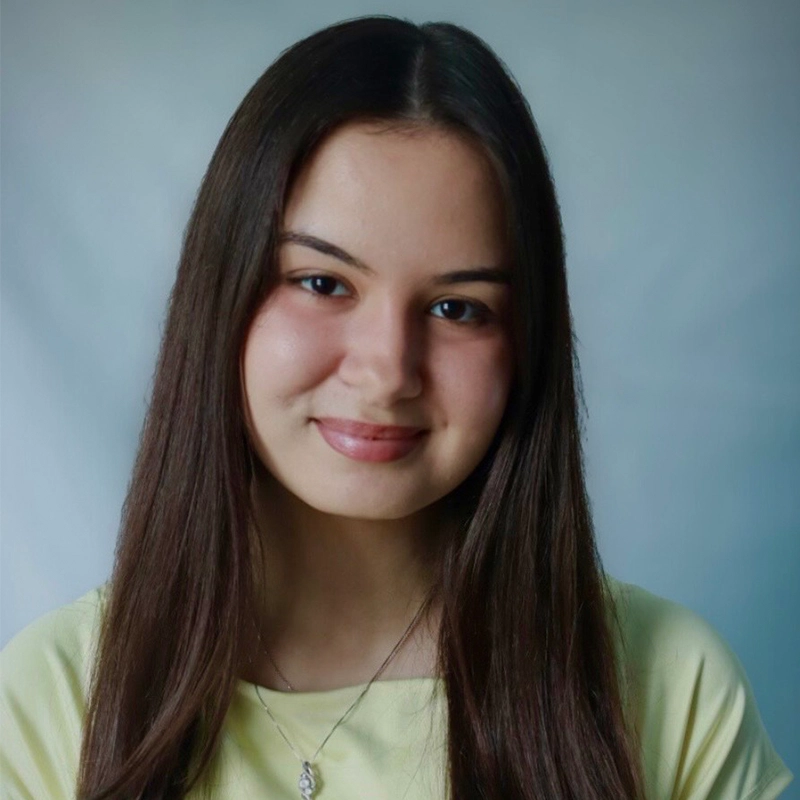 A young woman with straight, long brown hair and fair skin smiles gently at the camera. Theresa Landis wears a pale yellow top and a silver necklace with a small pendant. The background is a plain, light gradient.