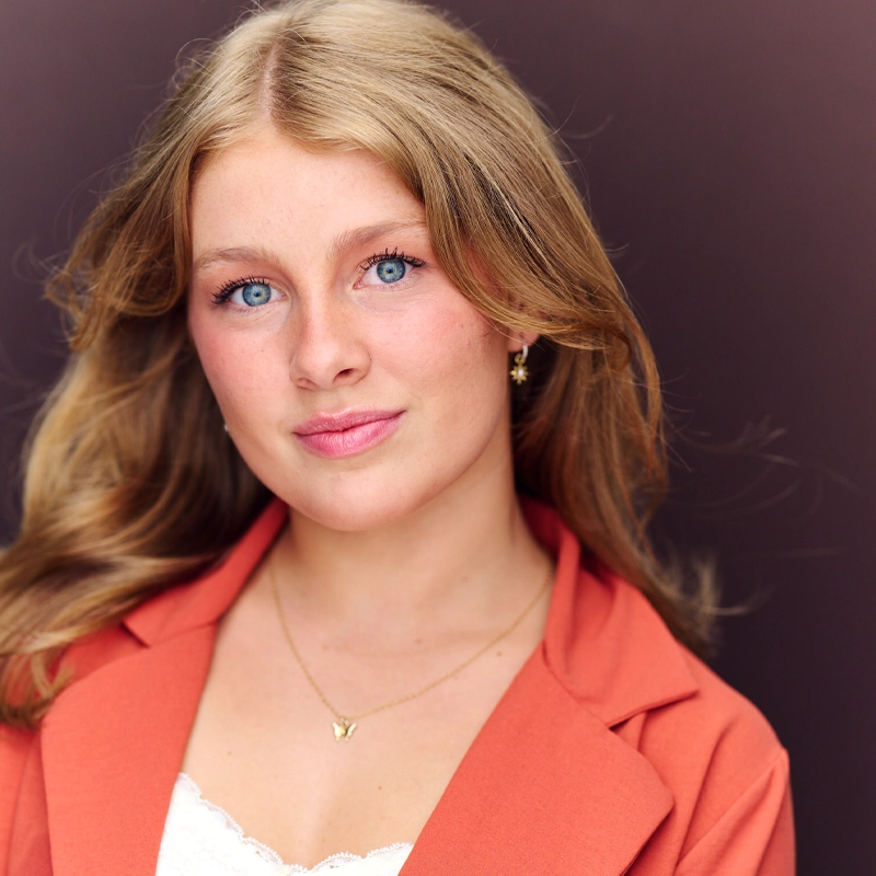 A young woman with long blonde hair and blue eyes faces the camera, wearing a coral blazer over a white lace top. She has minimal makeup, a gentle smile, gold earrings, and a delicate gold necklace. The background is a blurred, dark mauve shade.