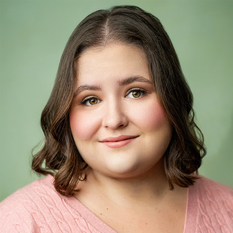 Grace Rundberg, a woman with wavy brown hair in a pink sweater, smiles softly at the camera. She has fair skin, hazel eyes, and natural makeup with rosy cheeks. The bright image features a soft, light green background and a friendly vibe.
