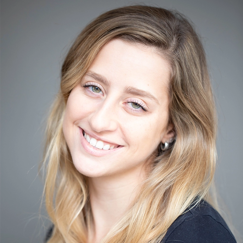 A young woman with long, wavy blonde hair smiles gently at the camera. She has light skin, blue eyes, and is wearing a black top. The background is plain and softly blurred, drawing focus to her friendly expression.