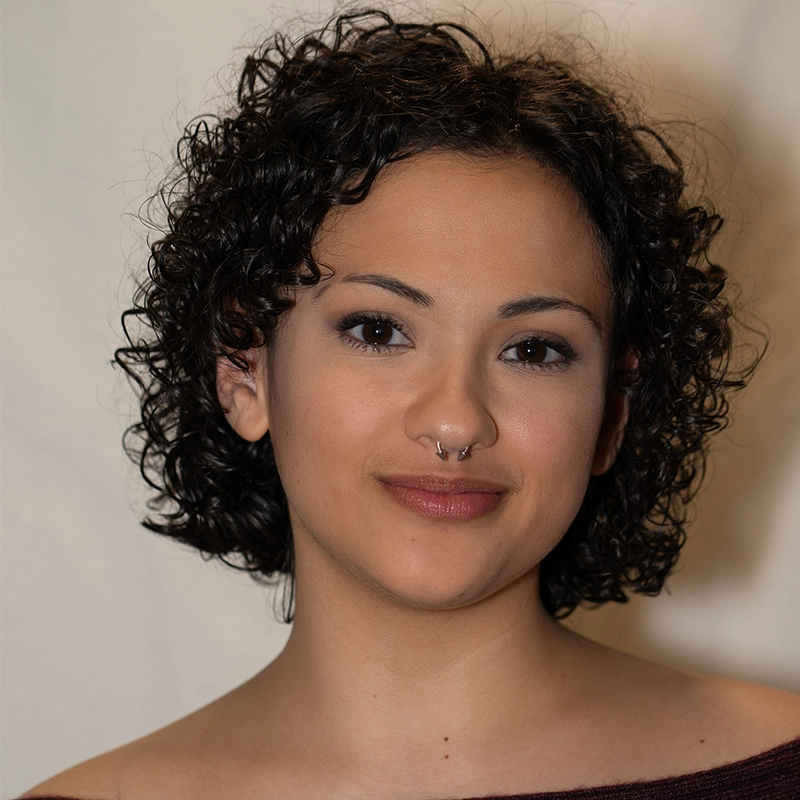 A person with short, curly black hair and light brown skin is facing the camera and smiling softly. Grace Rundberg has a septum piercing and wears subtle makeup. The plain, softly lit background draws focus to their face.