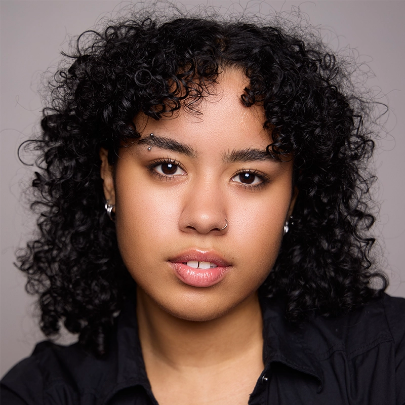 A person with medium brown skin, full lips, and black curly hair poses against a neutral background. With a nose ring, eyebrow piercing, and small hoop earrings, they wear a black collared shirt—exuding Grace Rundberg’s signature confidence at the camera.