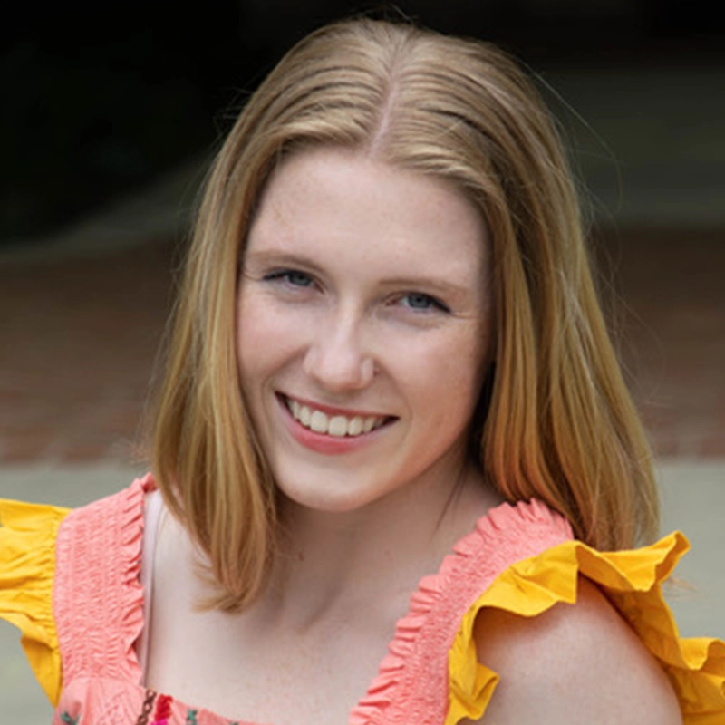 A young woman with straight, shoulder-length blonde hair smiles at the camera. She wears a light pink dress with yellow ruffled sleeves and a floral pattern. The background is softly blurred, showing an outdoor setting with a walkway and brick.