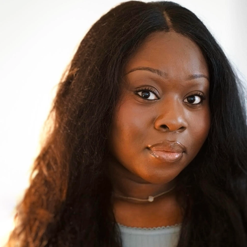 A woman with long, dark hair and smooth skin looks directly at the camera. She wears a light blue top and a delicate necklace with a small pendant. The background is softly lit and out of focus, emphasizing her calm, confident expression.