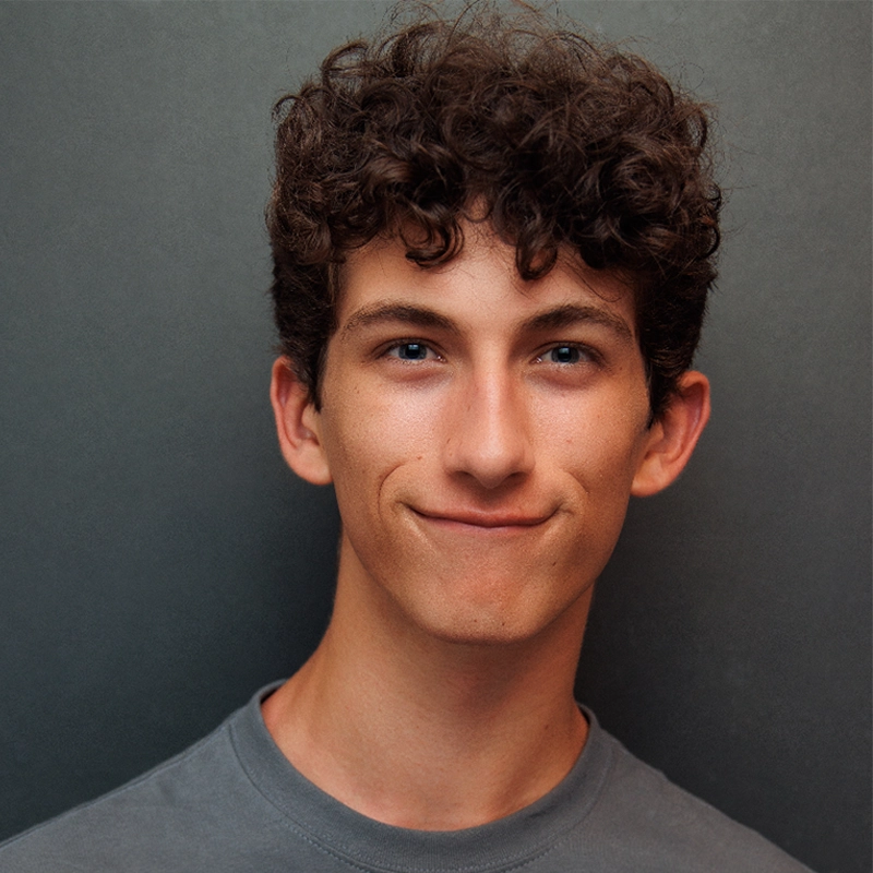 A young man with short, curly dark hair and blue eyes smiles softly at the camera. He wears a gray crew-neck shirt and stands before a plain, dark gray background, photographed in the style of Sylvie Friedman with even lighting highlighting his features.