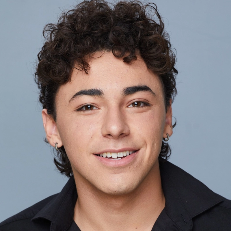Sylvie Friedman, a young person with short, curly dark hair, smiles warmly at the camera. They wear small hoop earrings and a black collared shirt. The plain light blue background gives the portrait a clean, professional look.