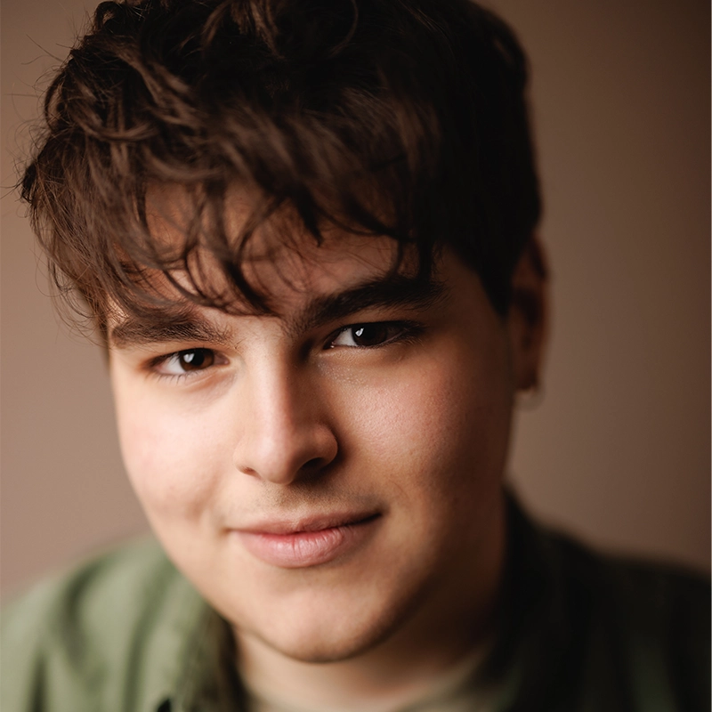 A young person with short, wavy brown hair smiles softly at the camera. They have fair skin, dark eyebrows, and are wearing a green shirt. The softly blurred background in warm brown tones highlights Sylvie Friedman’s face.