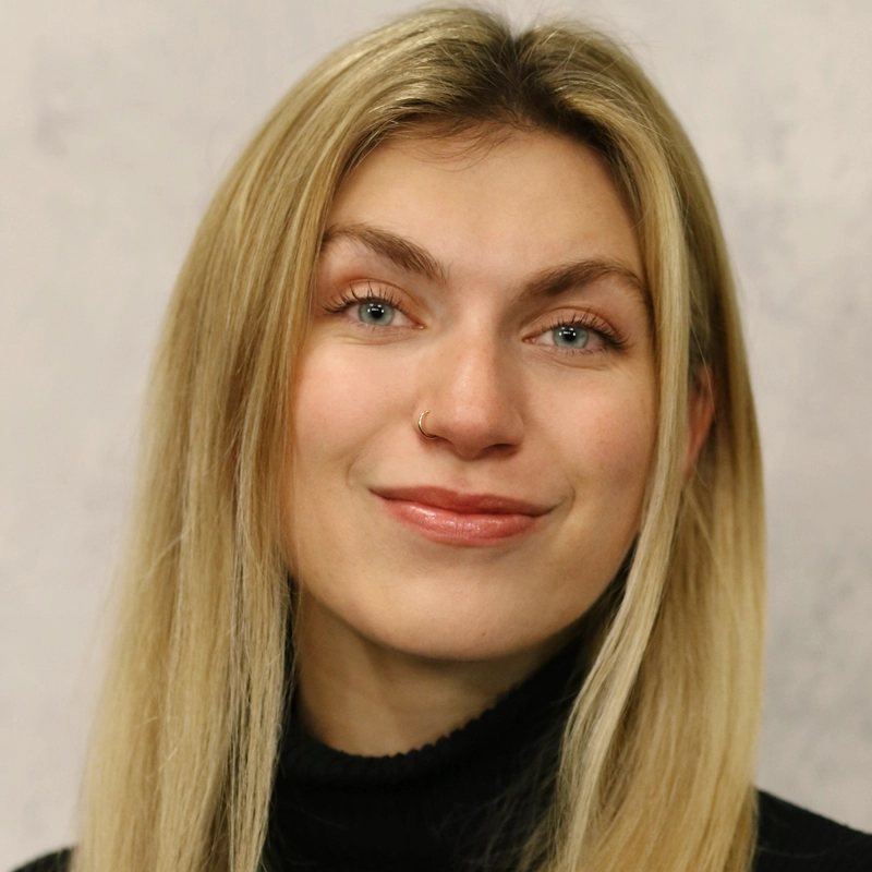 Meggie Ferguson, a young woman with long blonde hair, blue eyes, and a nose ring, smiles softly at the camera. She is wearing a black turtleneck sweater and is posed in front of a neutral, light-colored background.