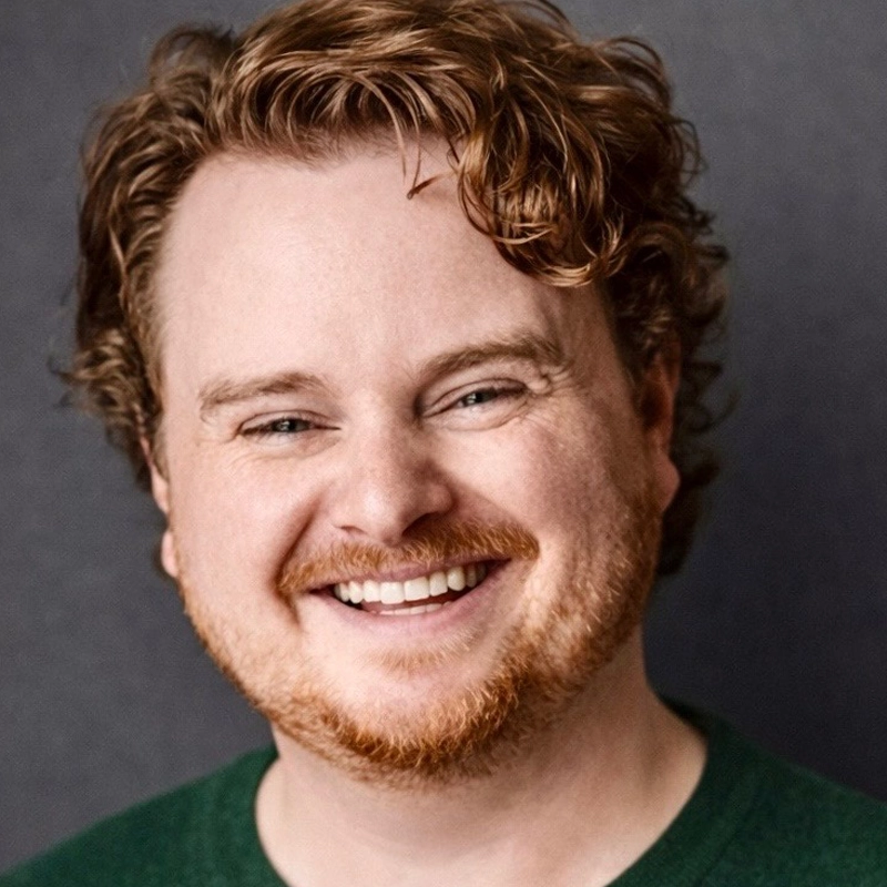 Jack Gereski, a man with fair skin, wavy red hair, and a short red beard, is smiling broadly. He wears a dark green sweater and poses against a plain, dark gray background. The well-lit photo highlights his cheerful expression.