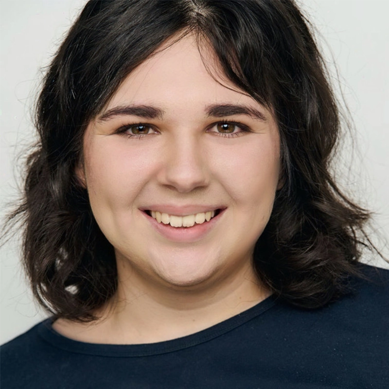 A person with wavy, dark brown hair and fair skin smiles at the camera. Wearing a navy blue top, Giuliana Gallone is posed against a plain light background, her expression friendly and approachable.