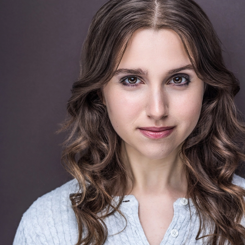 A young woman with wavy brown hair and fair skin looks directly at the camera, softly smiling. Jordan Greenberg is wearing a light blue textured shirt with buttons, set against a neutral brown background. The image is well-lit and sharply focused.