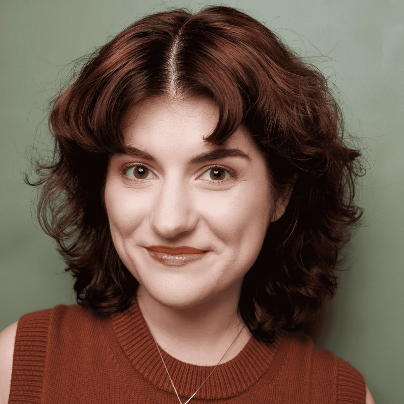 Taylor Greny, with wavy, shoulder-length brown hair, smiles softly in a sleeveless rust-colored top and necklace. Her natural makeup and friendly, approachable expression stand out against a plain green background.