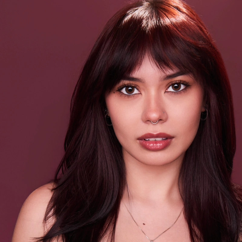 Woman with long dark brown hair and bangs, wearing natural makeup, a nose ring, hoop earrings, and a delicate necklace. She gazes directly at the camera against a solid maroon background, with soft lighting highlighting her features.