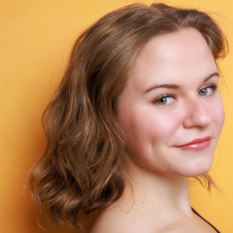 A young woman with light skin and wavy, shoulder-length brown hair smiles softly. Lydia Newman has light makeup, visible dimples, and wears a black strap. The background is a solid, warm yellow-orange color.