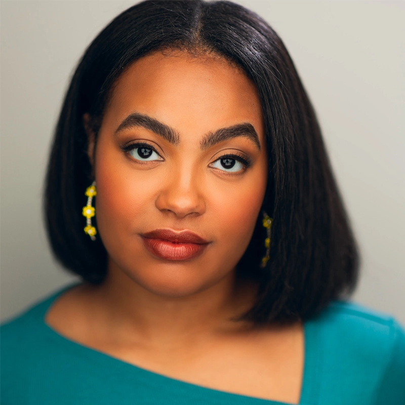 Nazarria Workman, a woman with medium brown skin and straight, shoulder-length dark hair, looks directly at the camera. She wears yellow dangling earrings and a teal top with an asymmetric neckline, exuding calm confidence against a plain background.