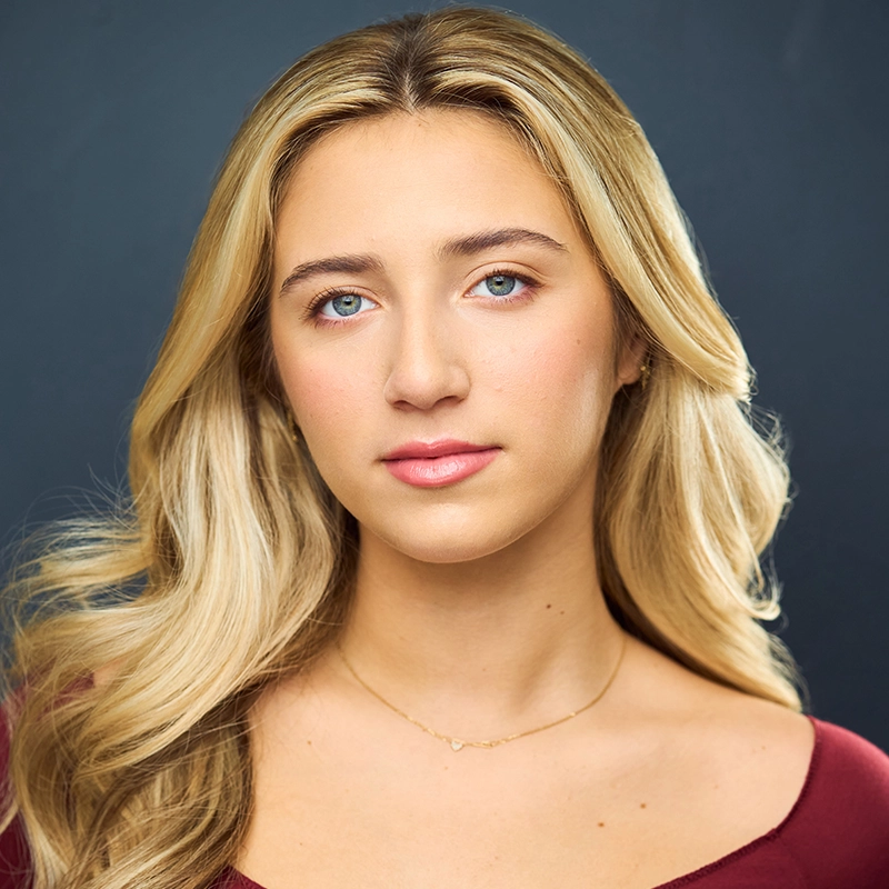 A young woman, Sylvie Friedman, with long, wavy blonde hair and blue eyes looks calmly at the camera. She wears a burgundy top and a thin gold necklace. The soft, blurred dark blue background highlights her natural features and neutral expression.