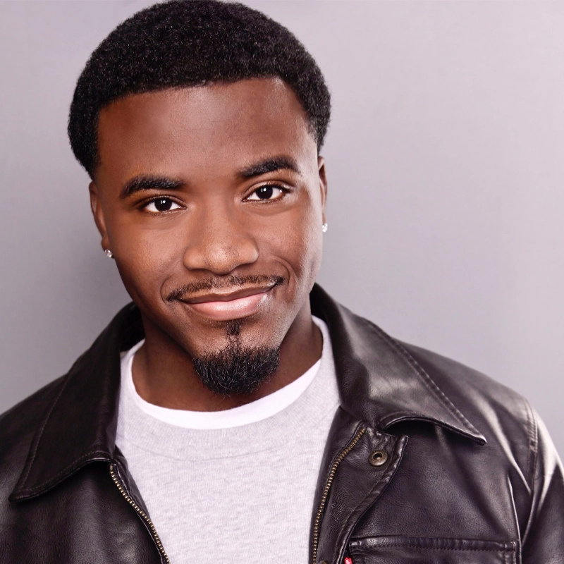 A young man with short, curly black hair and a trimmed goatee smiles warmly. He wears a black leather jacket over a light gray shirt, and small stud earrings. The background is plain and light gray, creating a simple and professional portrait.