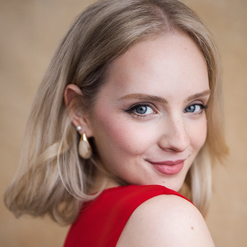 Brittany Smithson, a woman with fair skin, blue eyes, and shoulder-length blonde hair, smiles softly while glancing over her shoulder. She wears a red dress and gold teardrop-shaped earrings against a blurred light beige background.