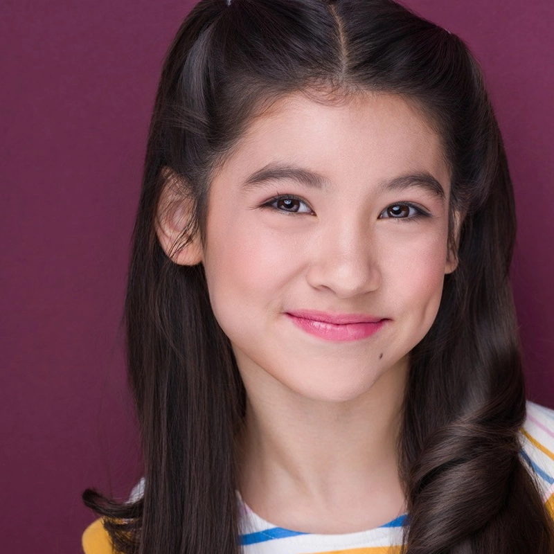 A young girl, Mari Southgate, with long dark brown hair styled in loose waves and half-up pigtails smiles warmly. She has light skin, deep brown eyes, and soft pink lipstick. She wears a white shirt with colorful stripes before a maroon background.