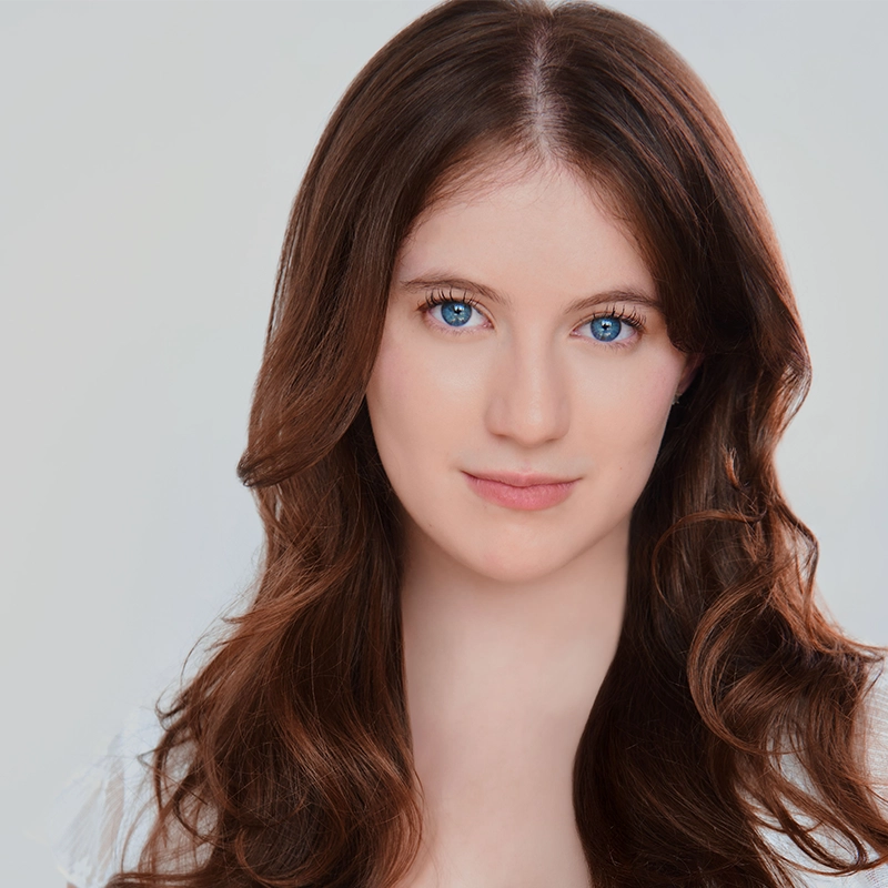 Sylvie Friedman, a young woman with long, wavy brown hair and striking blue eyes, looks directly at the camera. She has fair skin, a subtle smile, and wears a white top against a soft, light grey background for a clean portrait.