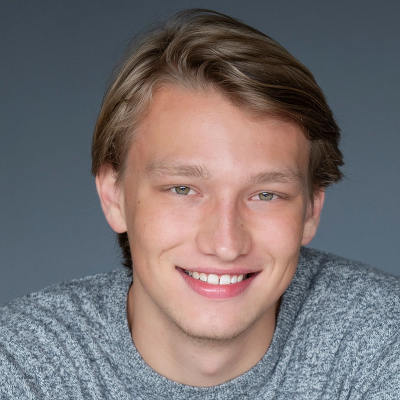 A young man with light skin, straight blonde hair parted to the side, and green eyes smiles warmly at the camera. He is wearing a grey knit sweater, styled in a look reminiscent of Sylvie Friedman, and is posed against a plain grey background.