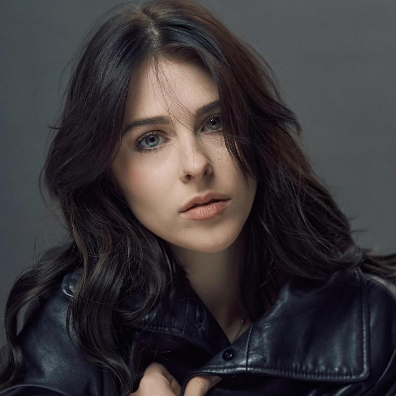 Ava Diane Tyson, a young woman with long wavy dark brown hair, stares at the camera. She has light skin, blue eyes, and natural makeup. She is wearing a black leather jacket and holds the collar with one hand. The background is neutral gray.