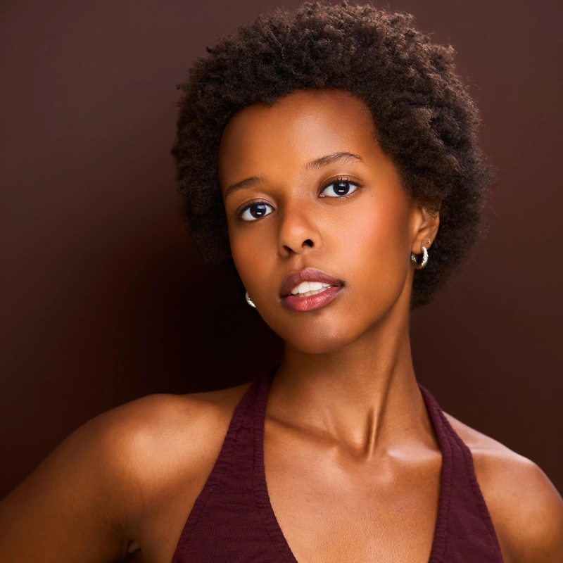 A young woman named Malaika, with short, curly hair, poses against a brown background. She has smooth, medium-brown skin, wears hoop earrings and a sleeveless burgundy top, gazing softly at the camera with a calm, confident expression.