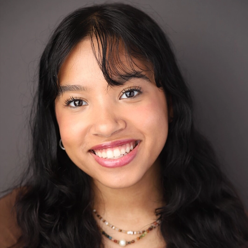 A young woman with long, wavy dark hair smiles at the camera. She has clear skin, wears natural makeup, and has layered necklaces. The background is plain and dark, keeping the focus on her face.