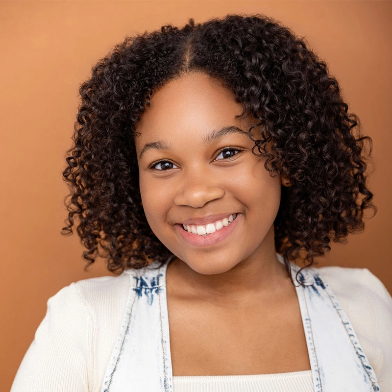 Ayvah Johnson, a young girl with curly dark hair, smiles warmly at the camera. She wears a cream-colored top with a light denim vest. The warm brown background and soft lighting highlight her cheerful expression and natural curls.