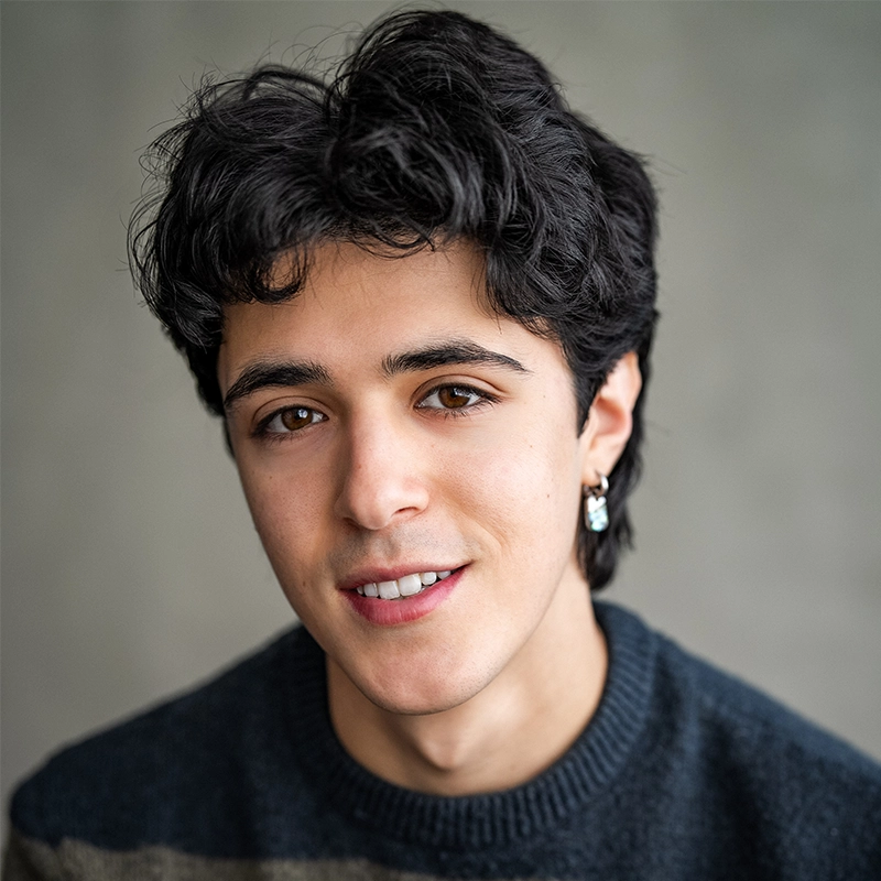Young person with medium-length wavy dark hair, wearing a dark sweater and a small earring, smiling gently at the camera. The background is softly blurred and gray, focusing attention on the person’s face.