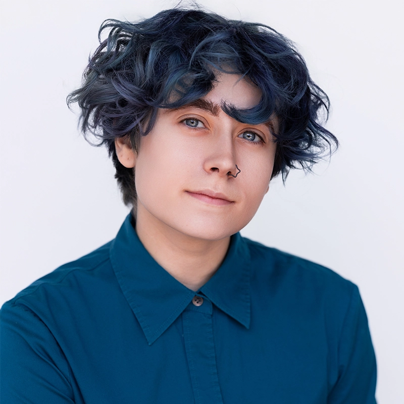 A person with short, wavy dark blue hair and a nose ring smiles softly at the camera. They wear a buttoned-up teal shirt and have light skin. The background is plain and white, creating a clean, simple portrait.