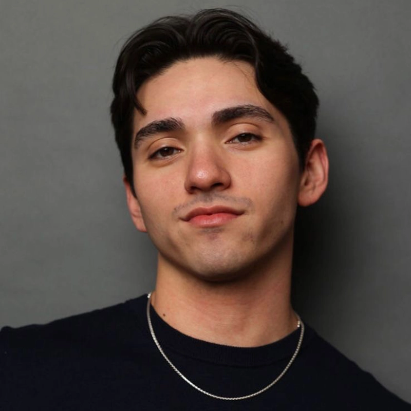 Diego Andres Rodriguez, a young man with short dark hair and light skin, poses against a plain gray background. He wears a dark crewneck shirt and a thin silver necklace, gazing at the camera with a slight smile and confident expression.