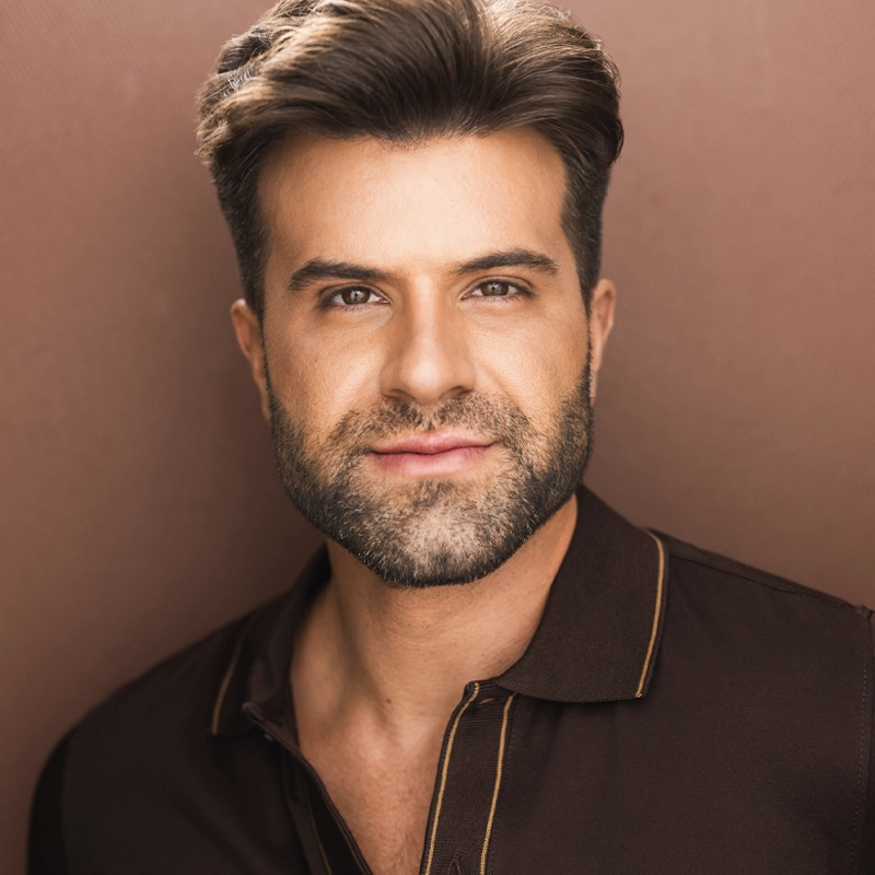 Danny Drewes, with styled brown hair and a trimmed beard, wears a dark brown collared shirt with yellow piping. He faces the camera and smiles slightly, standing against a plain light brown background under soft, even lighting.