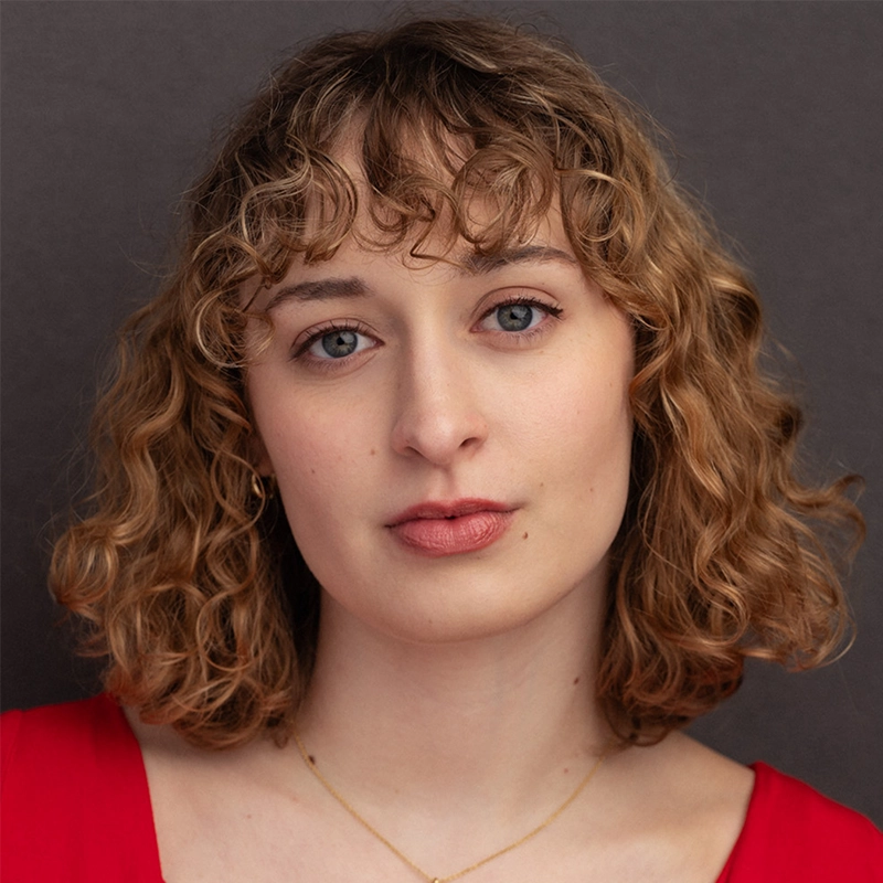 A young person with fair skin and short, curly light brown hair looks directly at the camera. They wear a red top and a delicate gold necklace. The background is plain and dark gray, and their expression is neutral and calm.