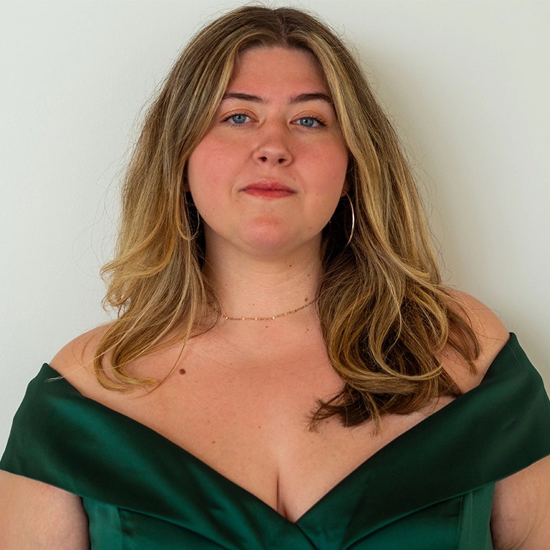 A woman with long, wavy blonde hair poses in front of a plain white background. She wears a deep green off-the-shoulder dress, large hoop earrings, and a delicate gold necklace. She looks directly at the camera with a neutral expression.