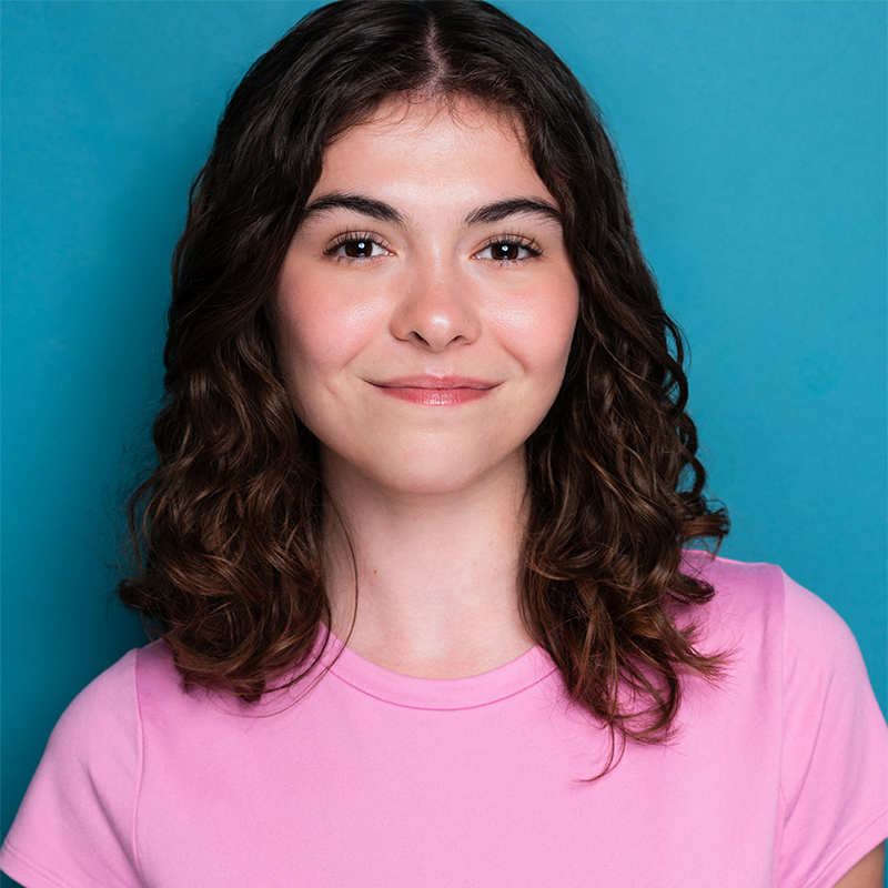 A young woman, Hailey Higgs, with medium-length curly brown hair smiles softly at the camera. She wears a light pink t-shirt and stands in front of a solid teal background. Her expression is calm and friendly.