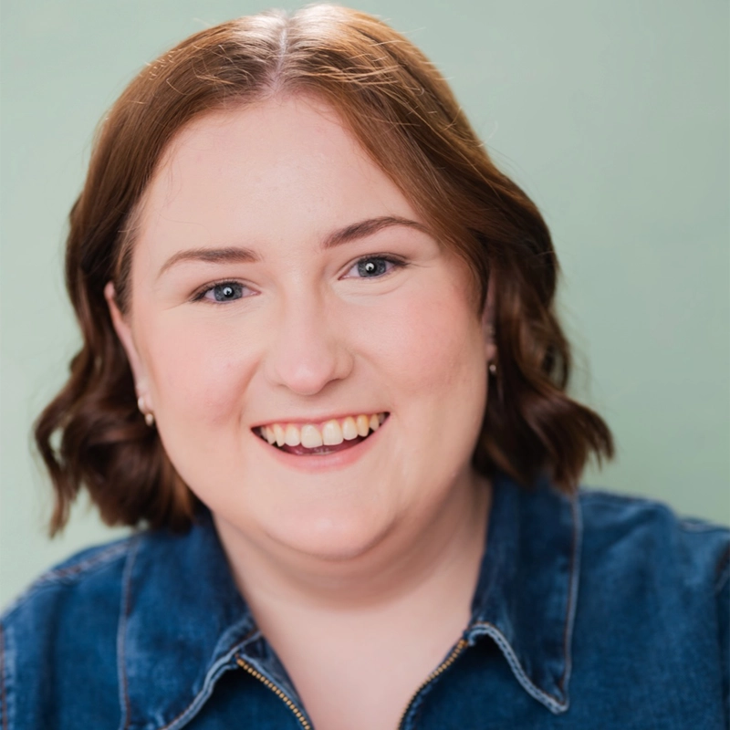 A woman with short, wavy, light brown hair smiles warmly at the camera. She has fair skin, blue eyes, and is wearing a denim shirt. The background is a plain light green color. The photo is well-lit and centered on her face.