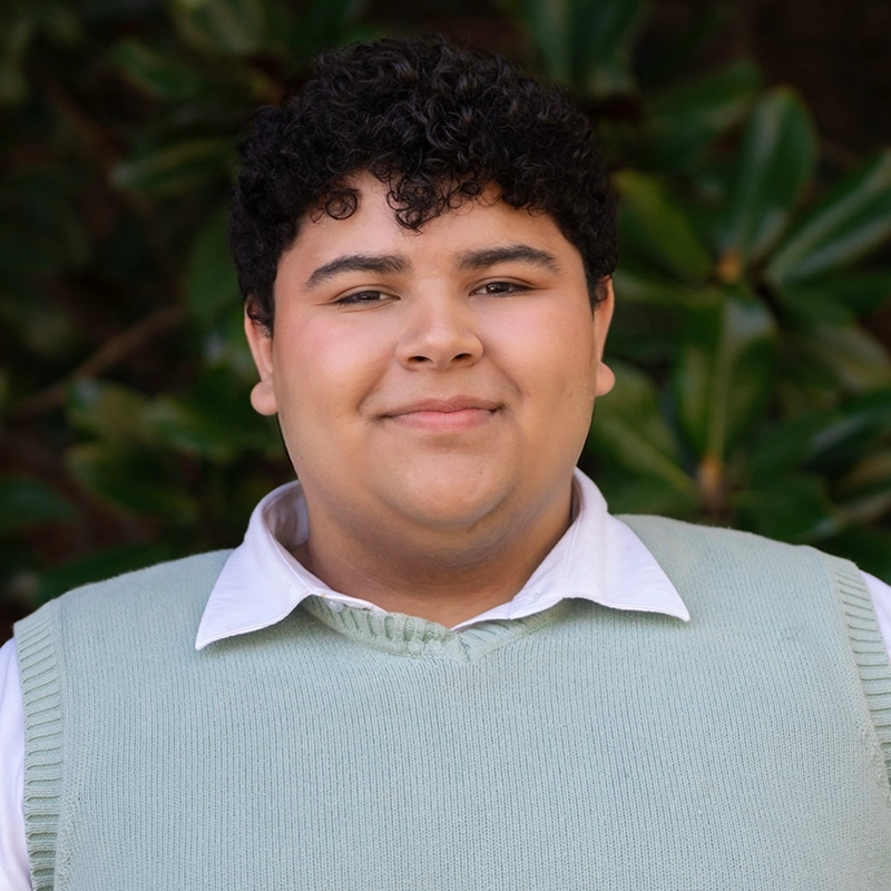A young person with curly dark hair smiles softly, wearing a white collared shirt under a light green sweater vest. The background is out of focus greenery, suggesting an outdoor setting.