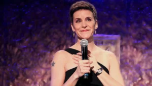 Jenn Colella, sporting a star tattoo on her upper arm, confidently holds a microphone on stage. Her short hair and sleeveless black top complement her hoop earrings. The backdrop features a blurred, textured pattern under purple lighting as she smiles, fully engaged with the audience.
