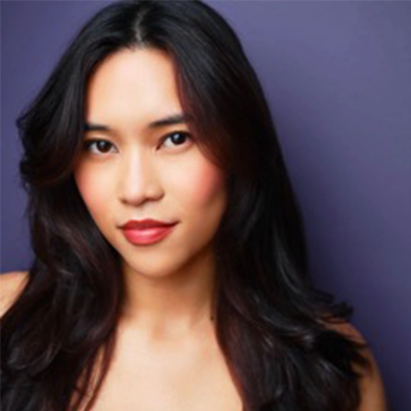 A woman with long, wavy dark hair and smooth skin poses in front of a purple background, gazing confidently at the camera. She has rosy cheeks, natural eye makeup, and wears a subtle red lipstick. Her shoulders are bare.